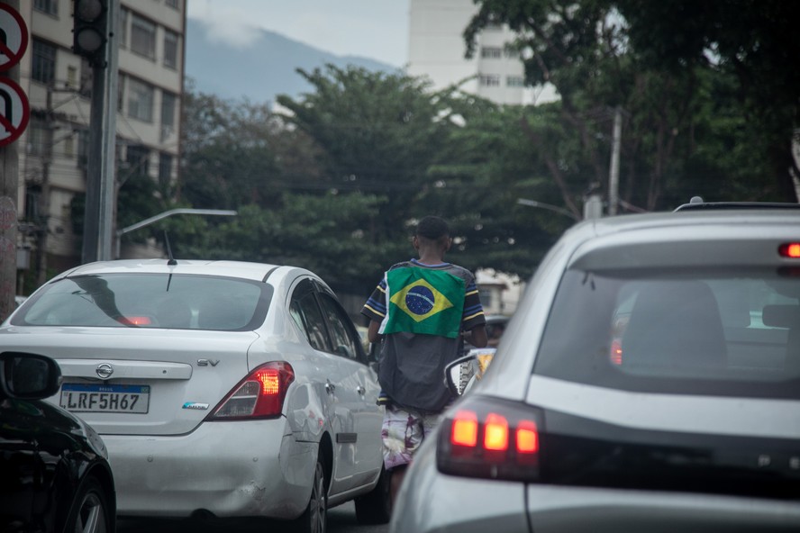 Vendedor de balas circula entre carros com bandeira do Brasil pendurada nas costas, na Barra da Tijuca, no Rio