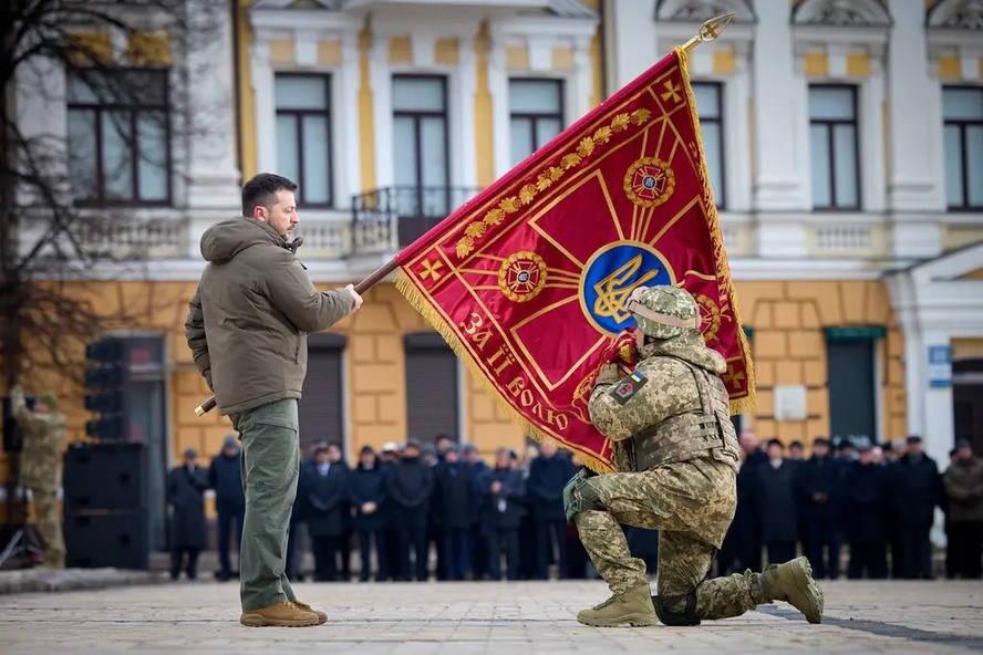 O presidente da Ucrânia, Volodymyr Zelensky, segura a bandeira de uma unidade militar enquanto um oficial a beija durante evento comemorativo em Kiev na sexta-feira