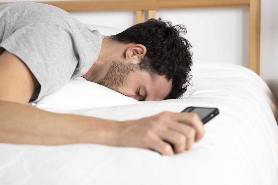 Homem deitado na cama com o rosto para baixo e o celular na mão