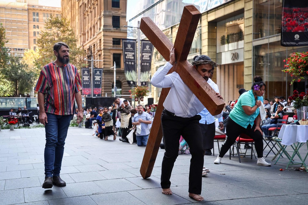 Na cidade de Sydney, na Austrália, um homem carrega uma cruzna encenação na Sexta-Feira Santa enquanto fiéis aobservam e se ajoelham — Foto: David Gray/ AFP