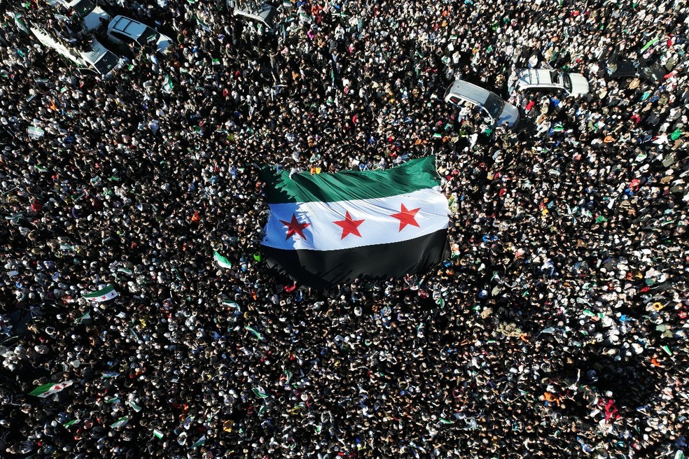 Sírios comemoram a queda do regime de Bashar al-Assad hasteando uma enorme bandeira da era da independência na Praça Umayyad, em Damasco, Síria, em 13 de dezembro de 2024 - Fotografia: Omar Haj Kaddour/AFP