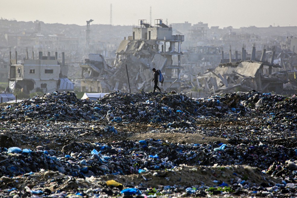 Um menino palestino procura material reciclável em um aterro sanitário tendo como pano de fundo prédios destruídos em Khan Yunis, no sul da Faixa de Gaza — Foto: Bashar Taleb / AFP