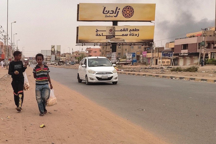 Duas crianças caminham com fumaça dos confrontos em Cartum, capital do Sudão, ao fundo