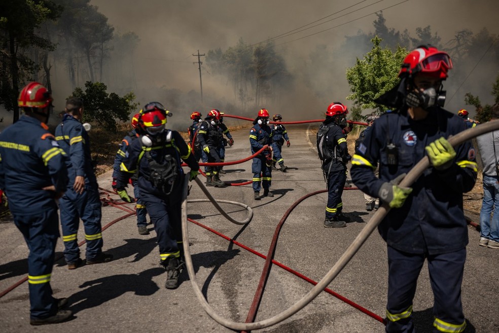 Bombeiros gregos seguram mangueiras durante operações de combate a um incêndio florestal na área de Porto Germeno, a cerca de 70 quilômetros (45 milhas) a noroeste de Atenas, na região da Beócia, em 2 de agosto de 2026 — Foto: ANGELOS TZORTZINIS / AFP