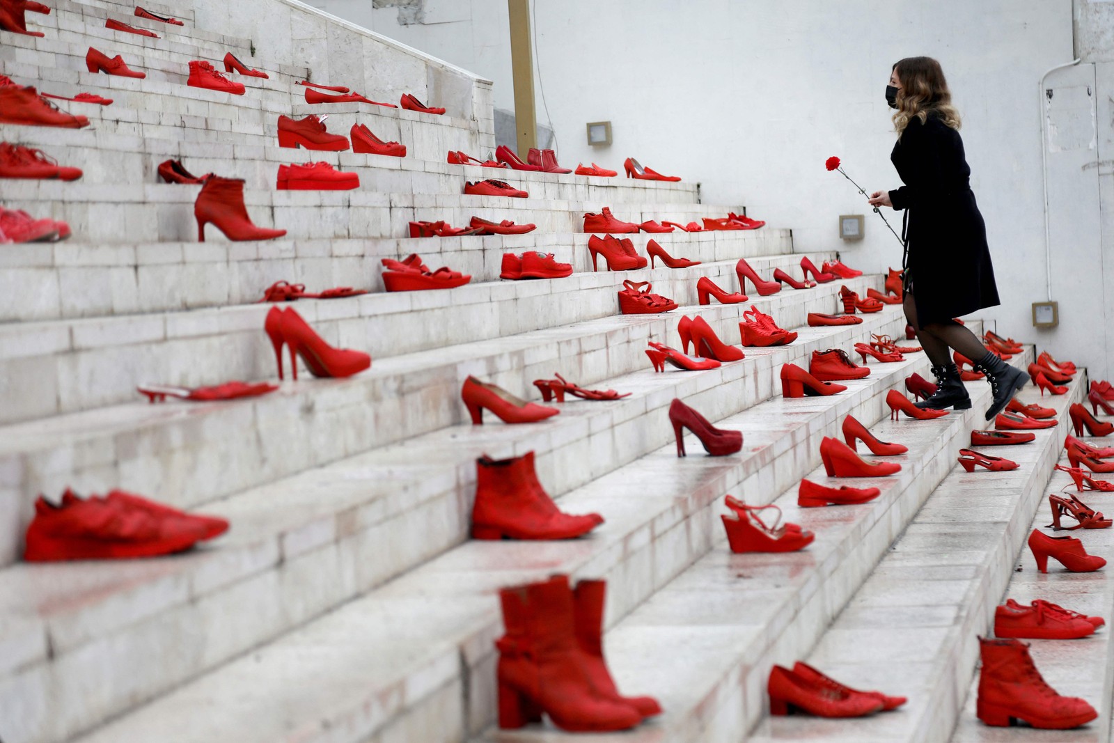 Mulher coloca uma flor vermelha em uma instalação de sapatos vermelhos femininos exibidos na escada, como um símbolo para denunciar a violência contra as mulheres, na praça principal de Durresi, em Tirana, AlbâniaAFP