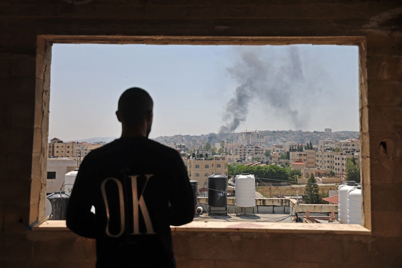 Homem palestino observa pela janela fumaça causada por ataques israelenses em Jenin — Foto: Jaafar Ashtiyeh/AFP
