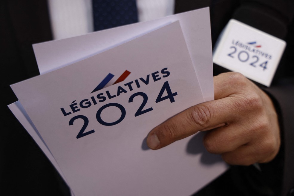 107530870-a-journalist-holds-cards-marked-legislatives-2024-during-the-election-night-of-the-governi ultradireita lidera com 34%, e esquerda avança; Macron pede 'união democrática' no 2° turno