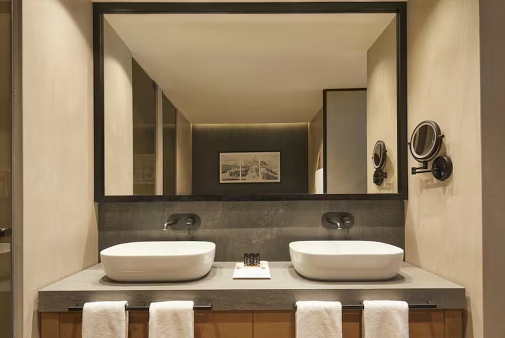 O banheiro privativo da suíte é equipado com chuveiro de hidromassagem, pias duplas e revestimentos de parede em tons neutros — Foto: Hotel MiM Baqueira