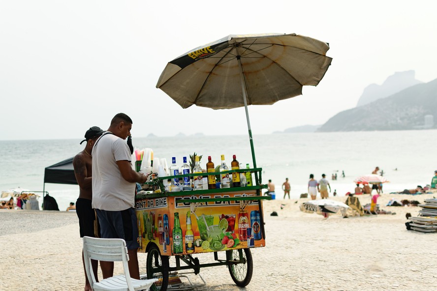 Vendedor de rua na praia do Rio vendendo bebidas sob um guarda-sol com fundo do oceano.