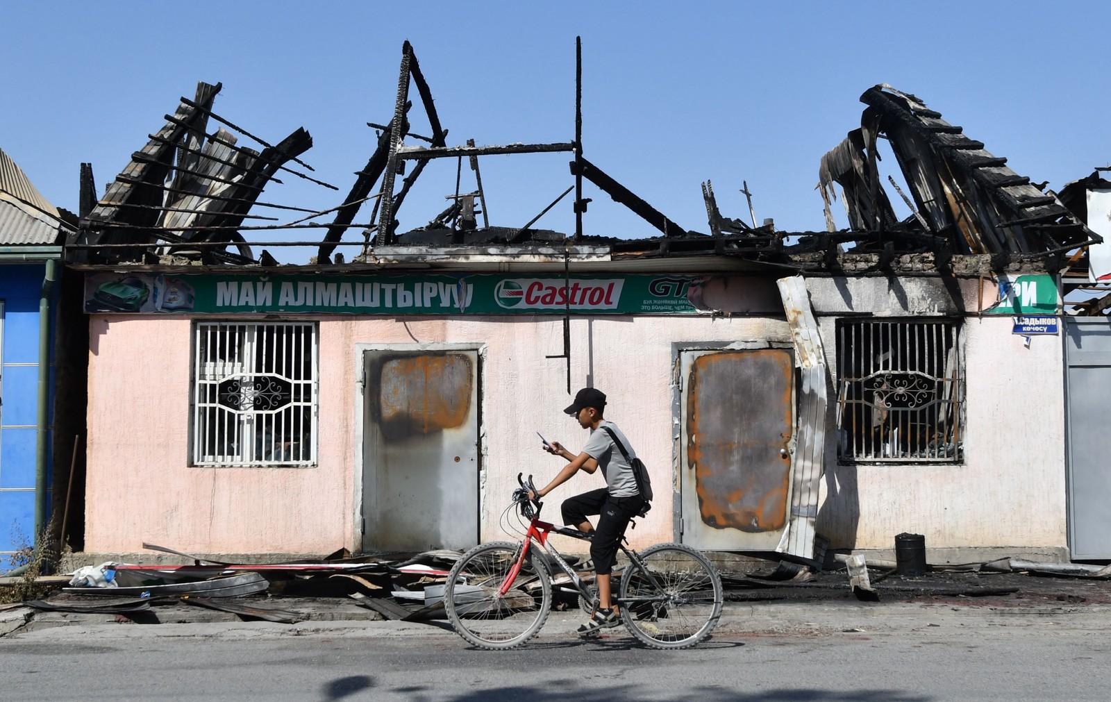 Jovem pedala uma bicicleta em frente a uma loja de óleo de automóveis queimada em Batken, a cerca de mil quilômetros de Bishkek, no Quirguistão — Foto: VYACHESLAV OSELEDKO / AFP