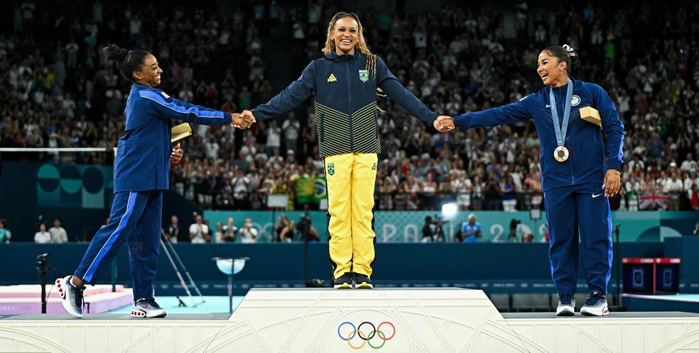 De mãos dadas com Simone Biles (prata) e Jordan Chiles (bronze), Rebeca Andrade se prepara para receber medalha de ouro no solo da ginástica artística — Foto: Gabriel BOUYS / AFP