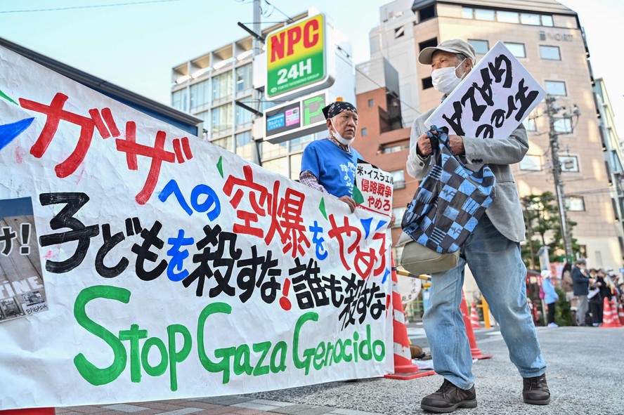 Homem com cartaz contra a guerra passa diante de manifestantes reunidos em Tóquio
