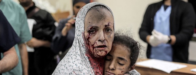 Mulher palestina ferida e coberta de poeira e sangue abraça uma menina também ferida no hospital após o bombardeio israelense de Khan Yunis, no sul da Faixa de Gaza, em 15 de novembro de 2023 — Foto: Belal KHALED / AFP