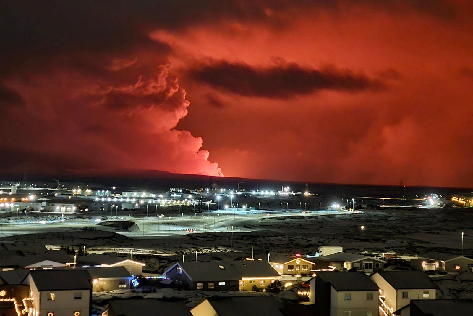 Casas na vila de Hafnarfjordur, enquanto a fuma&ccedil;a se eleva ao longe e a lava colore o c&eacute;u noturno de laranja &mdash; Foto: Oskar Grimur Kristjansson / AFP