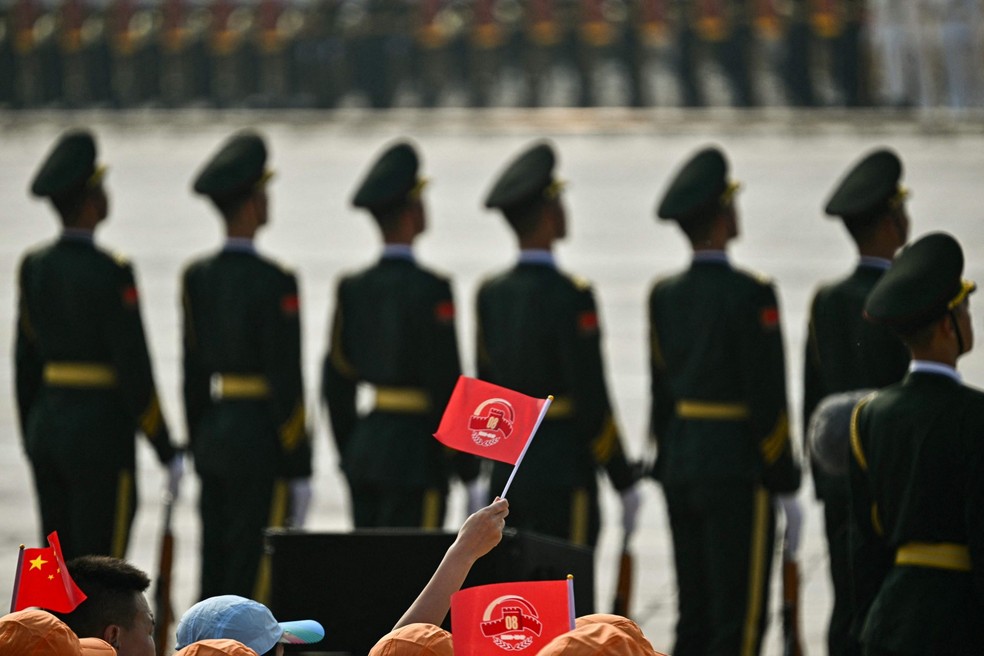 Um participante agita uma bandeira com o número'80' atrás de soldados chineses — Foto: Pedro Pardo / AFP