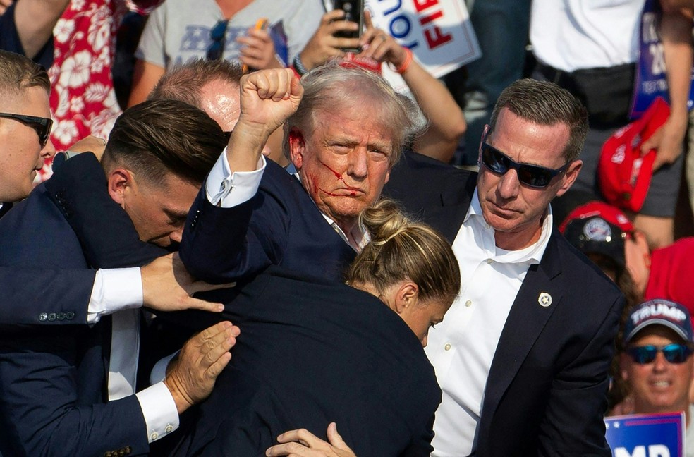 Donald Trump cercado por agentes do serviço secreto ao ser retirado do palco em um evento de campanha na Pensilvânia, em julho. O republicado foi atingido no ouvido em uma tentativa de assassinato por um homem armado no comício de campanha, em um acontecimento chocante que alimentou instabilidade antes das eleições presidenciais de 2024 nos EUA. O ex-presidente de 78 anos foi levado às pressas para fora do palco com sangue manchado no rosto após o tiroteio, enquanto o atirador e um espectador foram mortos e dois espectadores ficaram gravemente feridos — Foto: Rebecca DROKE / AFP