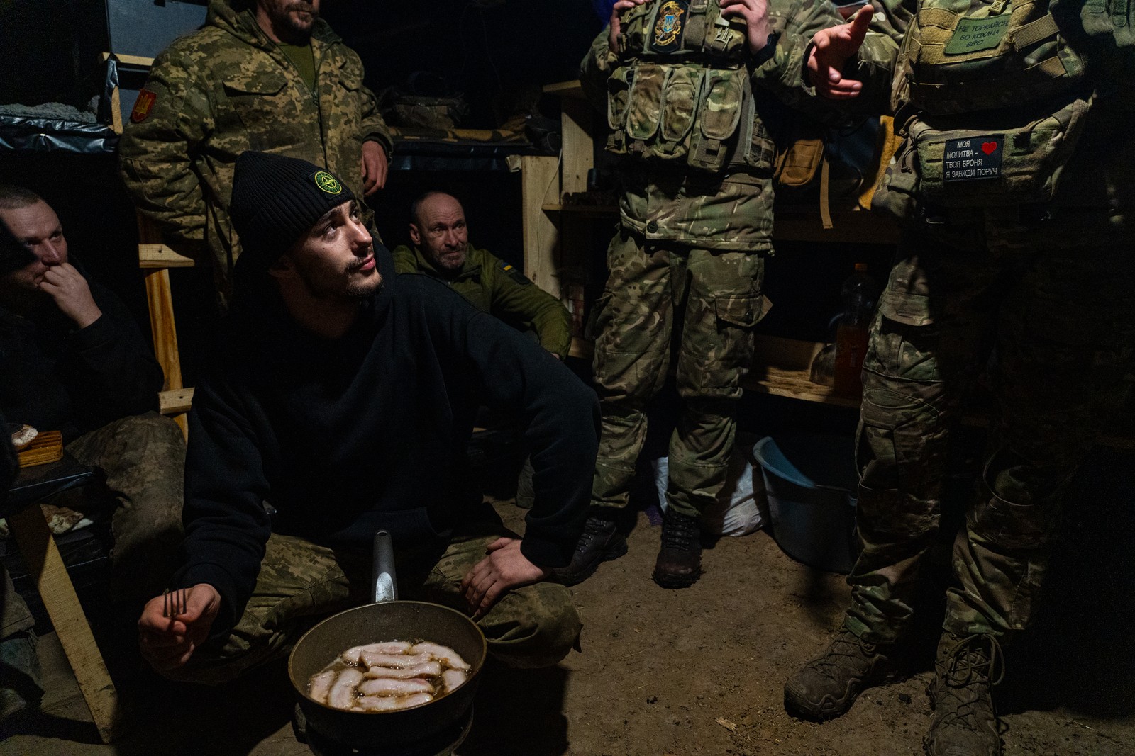 Soldados ucranianos fritam barriga de porco para comer como jantar em um bunker nas linhas de frente. Com os russos atacando e pouca munição soldados passam a maior parte do tempo em buracos como esse aguardando raras ordens para disparar contra o inimigo. Foto tirada em Kupiansk, Oblast de Kharkiv, Ucrânia. — Foto: Yan Boechat