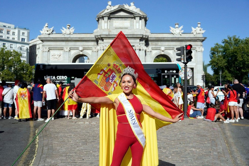 Uma mulher posa segurando uma bandeira da Espanha enquanto torcedores aguardam a passagem do desfile dos jogadores da seleção, um dia após a conquista do segundo título da Copa do Mundo com a vitória por 1 a 0 sobre a Argentina, na Puerta de Alcalá, em Madri, em 20 de julho de 2026.  Os campeões mundiais retornaram à capital espanhola para uma recepção de heróis, onde cerca de um milhão de torcedores eram esperados para saudá-los após uma noite de intensas comemorações por todo o país — Foto: Paul Hanna / AFP