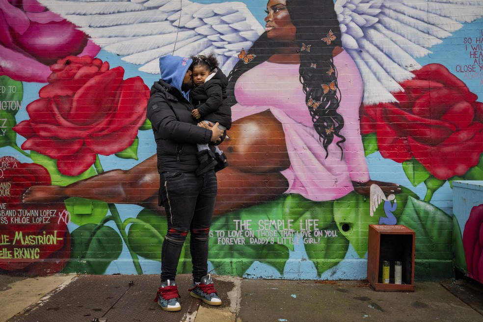 Juwan Lopez segura a filha, Khloe, diante de mural em homenagem à parceira, Sha-Asia Semple, morta após parto mal conduzido no Hospital Woodhull, símbolo da disparidade racial na mortalidade materna em NY — Foto: Desiree Rios/The New York Times